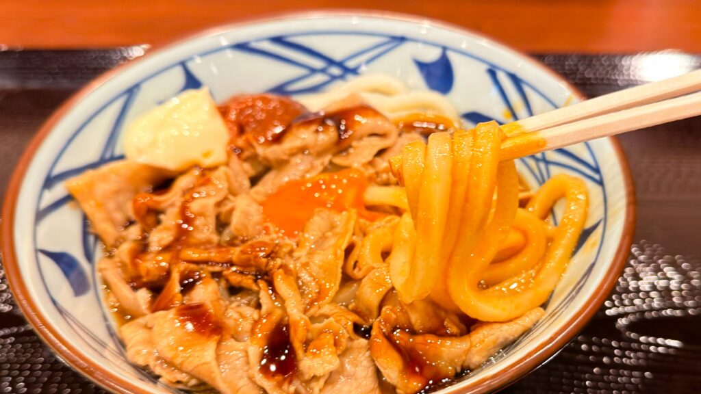 丸亀製麺_卵黄をつけた「こく旨豚玉ぶっかけうどん」の麺を箸で持っている様子