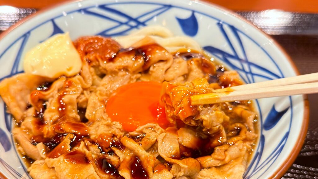 丸亀製麺_卵黄をつけた「こく旨豚玉ぶっかけうどん」の豚肉を箸で持っている様子